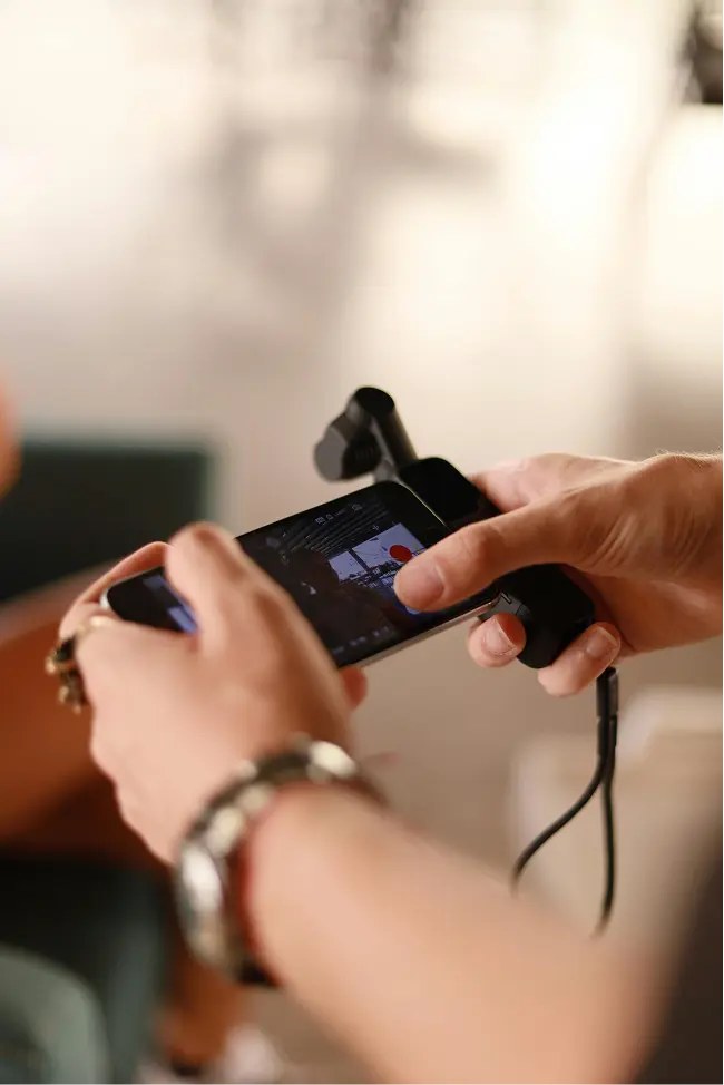 Hands hold a smartphone attached to a gimbal. The screen displays a recording interface with a red button. The setting is softly blurred and warmly lit.