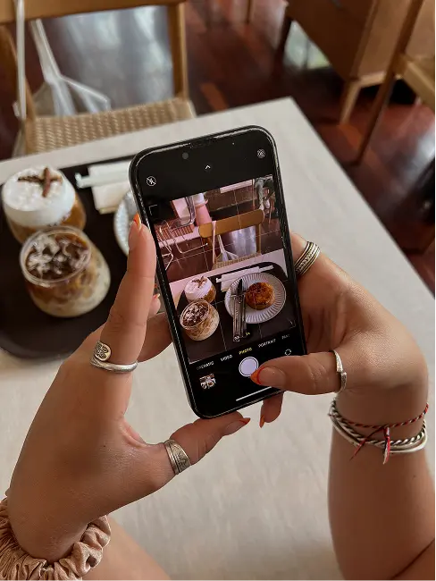 A content creator holding a phone capturing a photo of a table with iced coffee and dessert. The setting is cozy, conveying a relaxed and artistic mood.